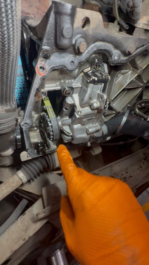 View from under a car, a gloved finger pointing at a small belt and sprocket on the engine, at AGR Performance, Stafford